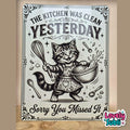 A rectangular decorative sign with a vintage-style black-and-white illustration of a cat in an apron holding a whisk and mixing bowl, with the text 'THE KITCHEN WAS CLEAN YESTERDAY Sorry You Missed It.' The sign is photographed standing upright against a beige wall on a wooden surface.