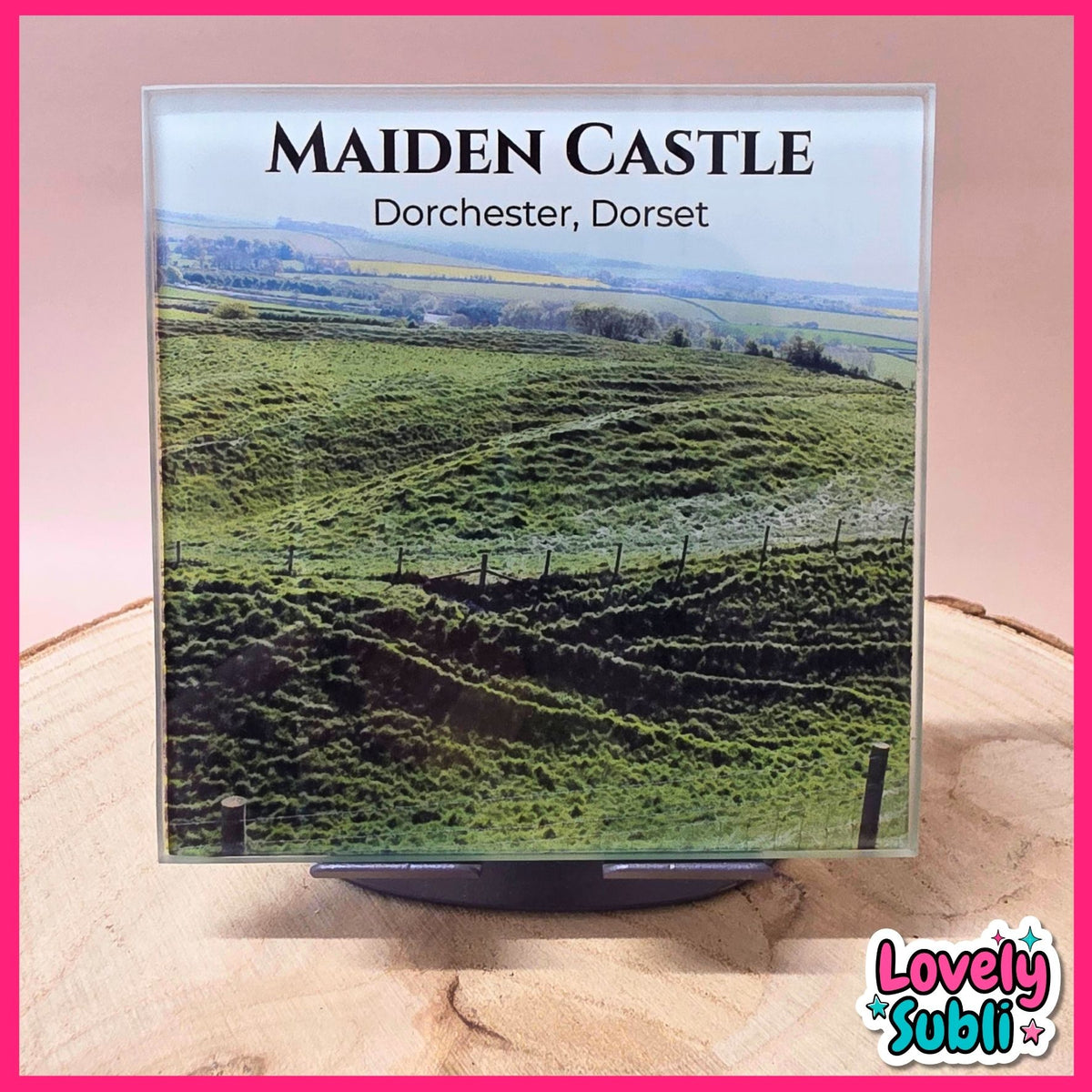 A square product display featuring a printed scenic image of rolling green hills with the text 'MAIDEN CASTLE Dorchester, Dorset' at the top, mounted on a black stand against a pink background with a wood slice surface.