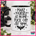A white ceramic mug with a black handle and black rim, featuring the text 'MAKE YOURSELF AT HOME FUCK OFF BY 9PM' in black lettering with a black moth graphic below. The mug is set on a marble surface with a blurred background containing candles and plants.