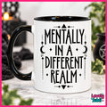 A white ceramic mug with a black handle and black interior, printed with the phrase 'MENTALLY IN A DIFFERENT REALM' in black lettering with celestial and geometric motifs. The mug is shown on a marble surface with a blurred cozy background featuring candles and plants.