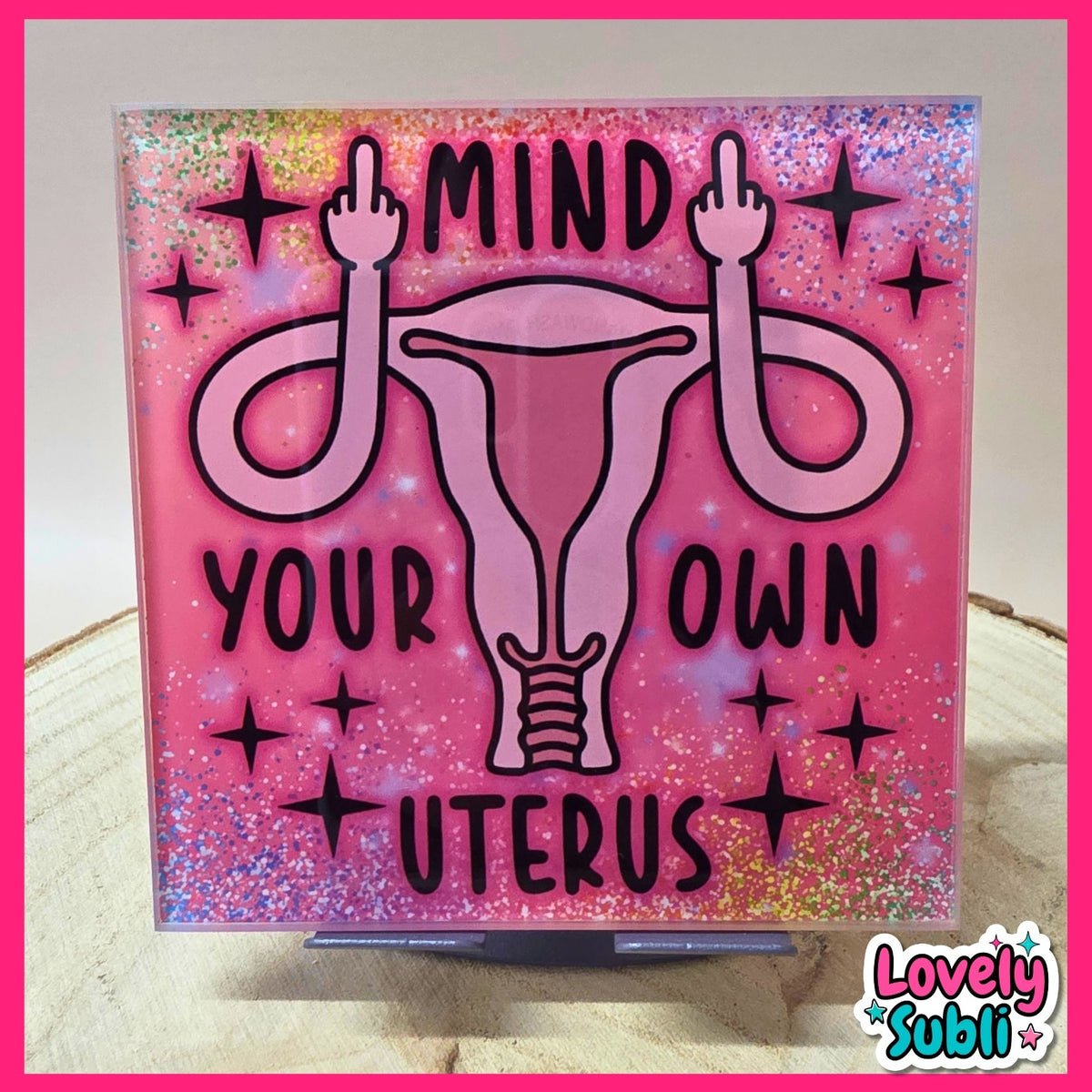 A square acrylic art plaque featuring a pink uterus graphic with the words 'MIND YOUR OWN UTERUS' in black text, surrounded by pink glitter. The plaque is displayed on a small stand on a wooden surface.