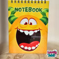 A spiral-bound notebook with a bright yellow cover featuring a cartoon monster face with green horns and the word 'NOTEBOOK' at the top. The notebook is standing on a wooden surface against a neutral beige background, with a 'Lovely Subli' watermark in the bottom right corner.