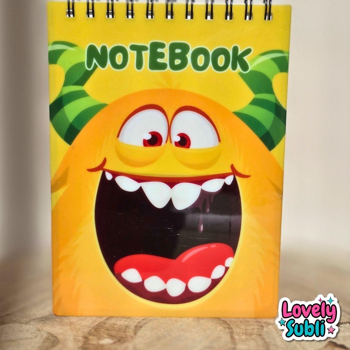 A spiral-bound notebook with a bright yellow cover featuring a cartoon monster face with green horns and the word 'NOTEBOOK' at the top. The notebook is standing on a wooden surface against a neutral beige background, with a 'Lovely Subli' watermark in the bottom right corner.