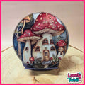 A hand-painted glass snow globe featuring whimsical fairy tale mushroom houses with red and white spotted caps, set on a small blue stand. The globe is displayed on a light wood slice against a beige background, framed by a pink border with a 'Lovely Subli' brand logo in the bottom right corner.