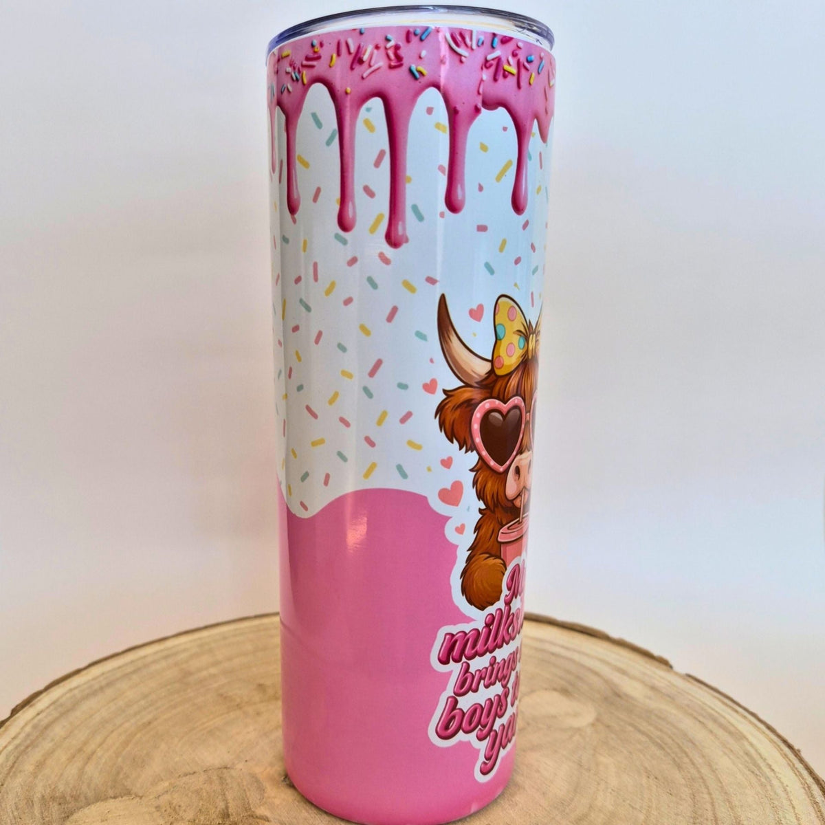 A tall insulated tumbler with a pink and white donut-themed design, featuring dripping pink glaze, colorful sprinkles, a cartoon cow wearing heart-shaped sunglasses, and partial text on a light wood slice background.