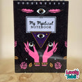 A spiral-bound notebook with a black cover featuring pink feathers, a central eye illustration, the text 'My Mystical NOTEBOOK' on a white label, pink hands holding three eyes, and pink roses. The notebook is photographed standing upright on a wooden surface against a beige background, with a 'Lovely Subli' watermark in the bottom right corner.