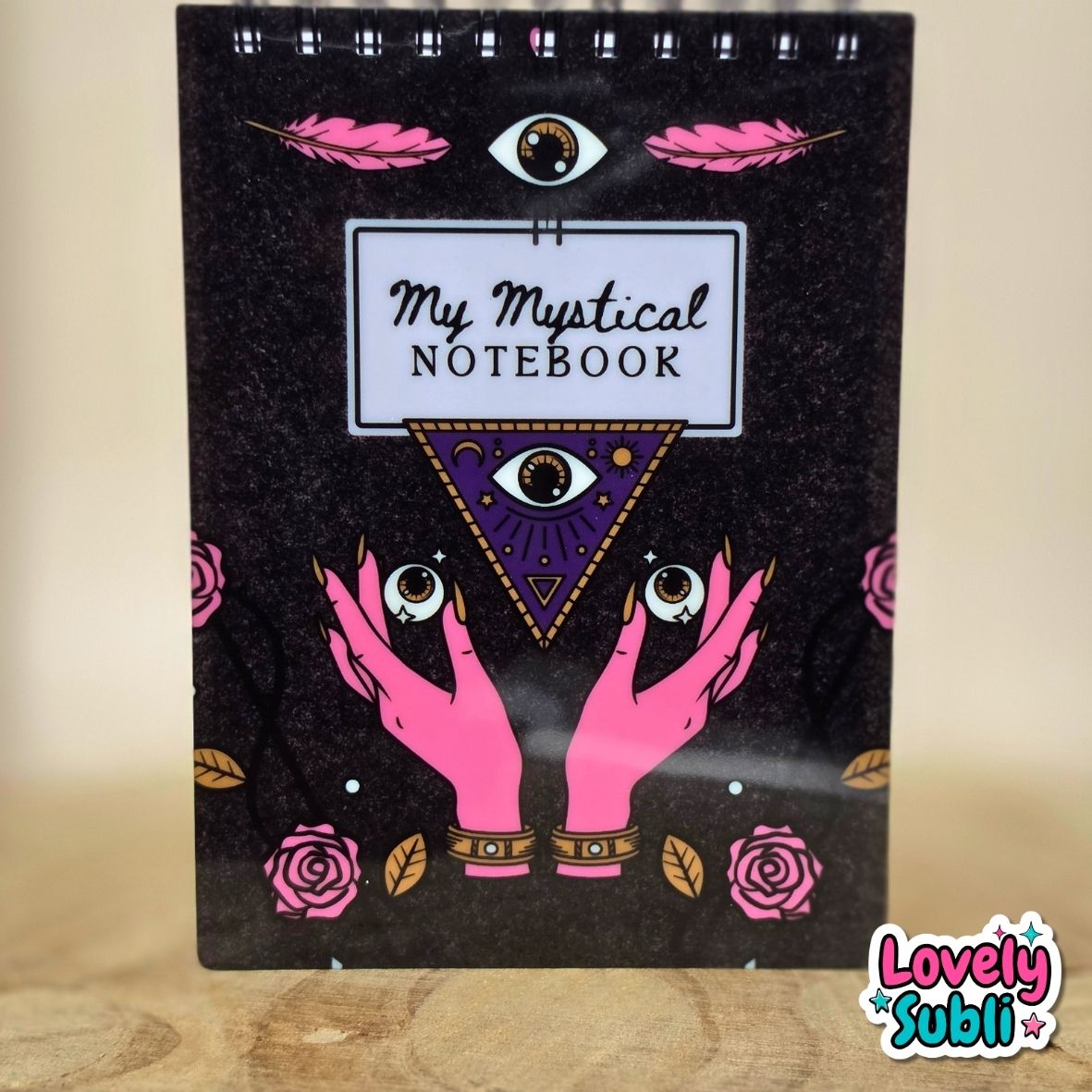 A spiral-bound notebook with a black cover featuring pink feathers, a central eye illustration, the text 'My Mystical NOTEBOOK' on a white label, pink hands holding three eyes, and pink roses. The notebook is photographed standing upright on a wooden surface against a beige background, with a 'Lovely Subli' watermark in the bottom right corner.