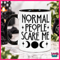 A white ceramic mug with a black handle and black interior, printed with the phrase 'NORMAL PEOPLE SCARE ME' in black lettering above moon and star symbols. The mug is photographed on a marble-like surface with blurred dark decorative objects and greenery in the background.