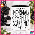 A white ceramic mug with a black handle and black interior, printed with the phrase 'NORMAL PEOPLE SCARE ME' in a gothic-style black font, surrounded by decorative symbols. The mug is set on a marble surface with a blurred background containing candles and plants.