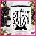 A white ceramic mug with a black handle and black interior, printed with the phrase 'NOT TODAY SATAN' above a black cat face. The mug is shown on a marble-like surface with a blurred dark background and greenery.