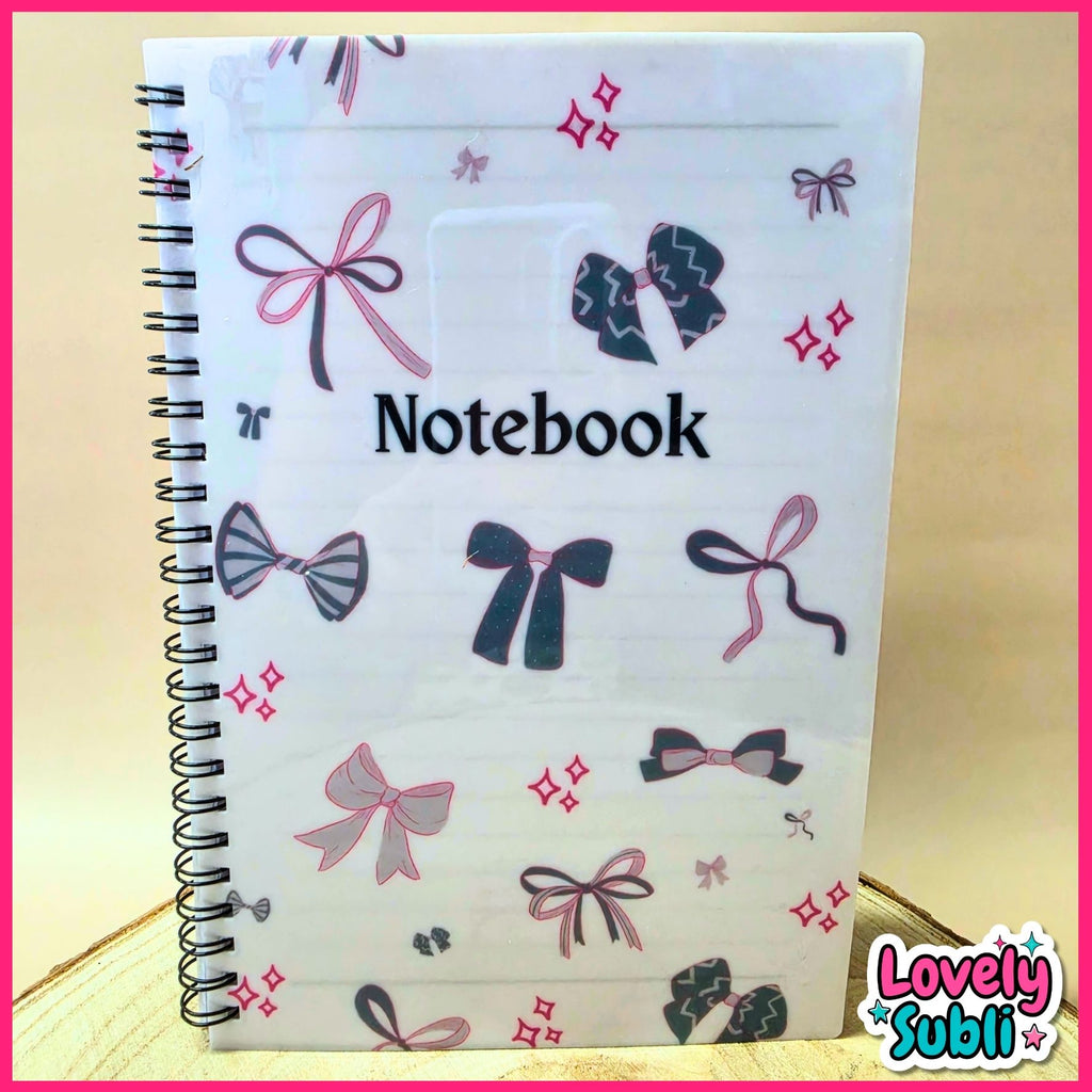 A white spiral-bound notebook with a cover decorated with pink, purple, and black bow illustrations and the word 'Notebook' printed in black. The notebook is photographed on a light wood surface with a bright pink border around the image.