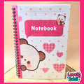 A spiral-bound notebook with a cute cartoon bear face and pink hearts on the cover, featuring a pink speech-bubble label that says 'Notebook' and a small bear holding a heart. The notebook is photographed on a light wood surface with a bright pink border around the image.