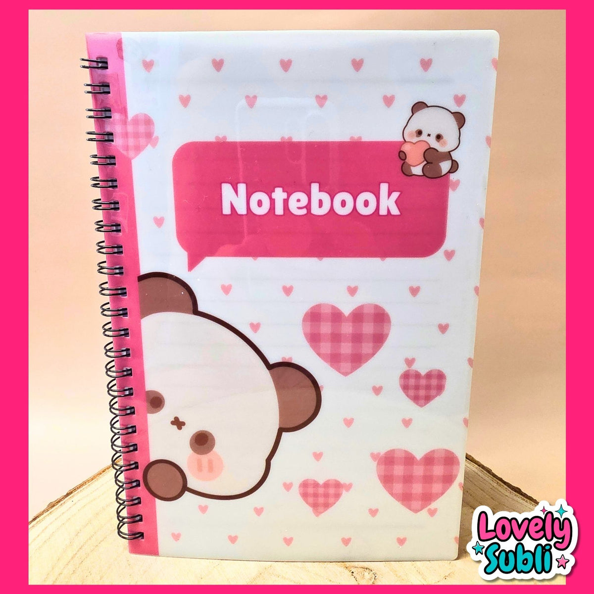 A spiral-bound notebook with a cute cartoon bear face and pink hearts on the cover, featuring a pink speech-bubble label that says 'Notebook' and a small bear holding a heart. The notebook is photographed on a light wood surface with a bright pink border around the image.