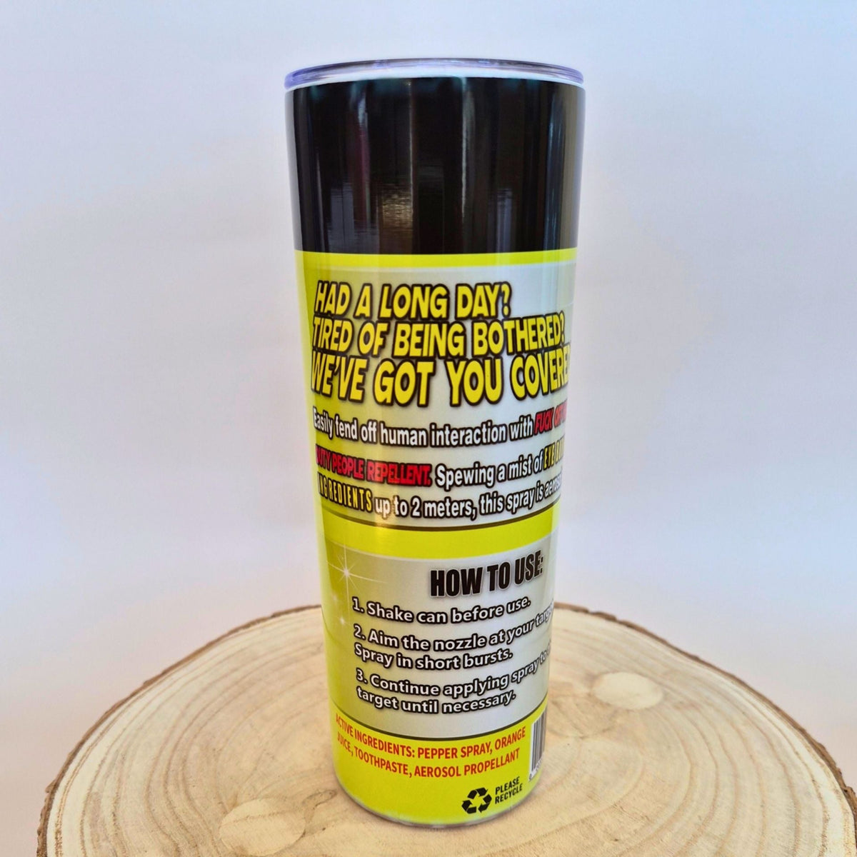 A novelty pepper spray can with a black top and a bright yellow label featuring humorous text and usage instructions, displayed on a circular wood slice against a light gray background.