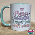 A white ceramic mug with a light blue handle and interior, featuring the printed text 'Please assume I want to be left alone' in pastel pink, purple, and green lettering with heart and star accents. The mug sits on a wooden surface against a neutral beige background, with a 'Lovely Subli' watermark in the bottom right corner.