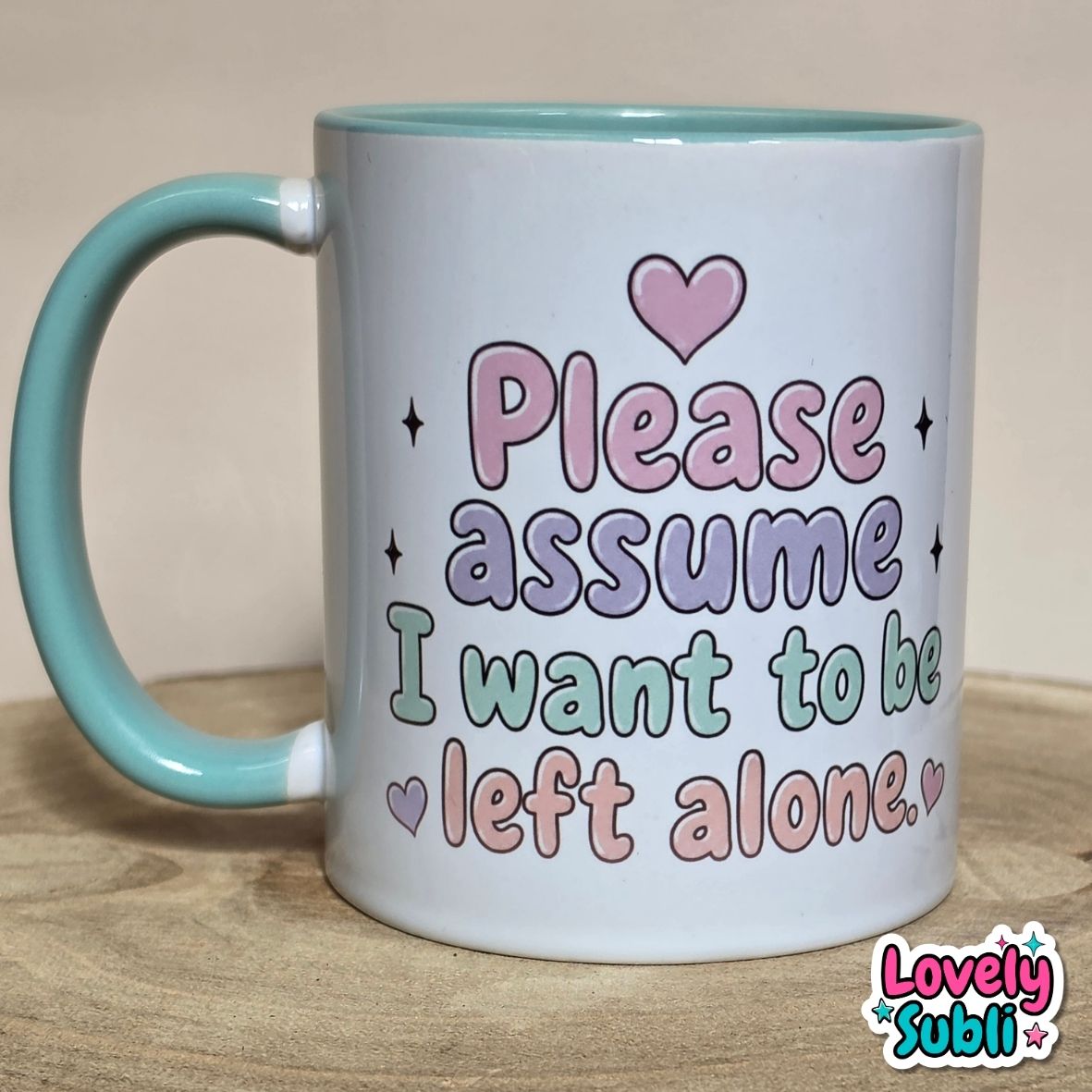 A white ceramic mug with a light blue handle and interior, featuring the printed text 'Please assume I want to be left alone' in pastel pink, purple, and green lettering with heart and star accents. The mug sits on a wooden surface against a neutral beige background, with a 'Lovely Subli' watermark in the bottom right corner.