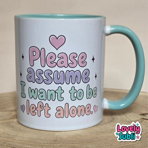A white ceramic mug with a light teal handle and interior, featuring the text 'Please assume I want to be left alone.' in pastel pink, purple, and green lettering with small heart and star accents.