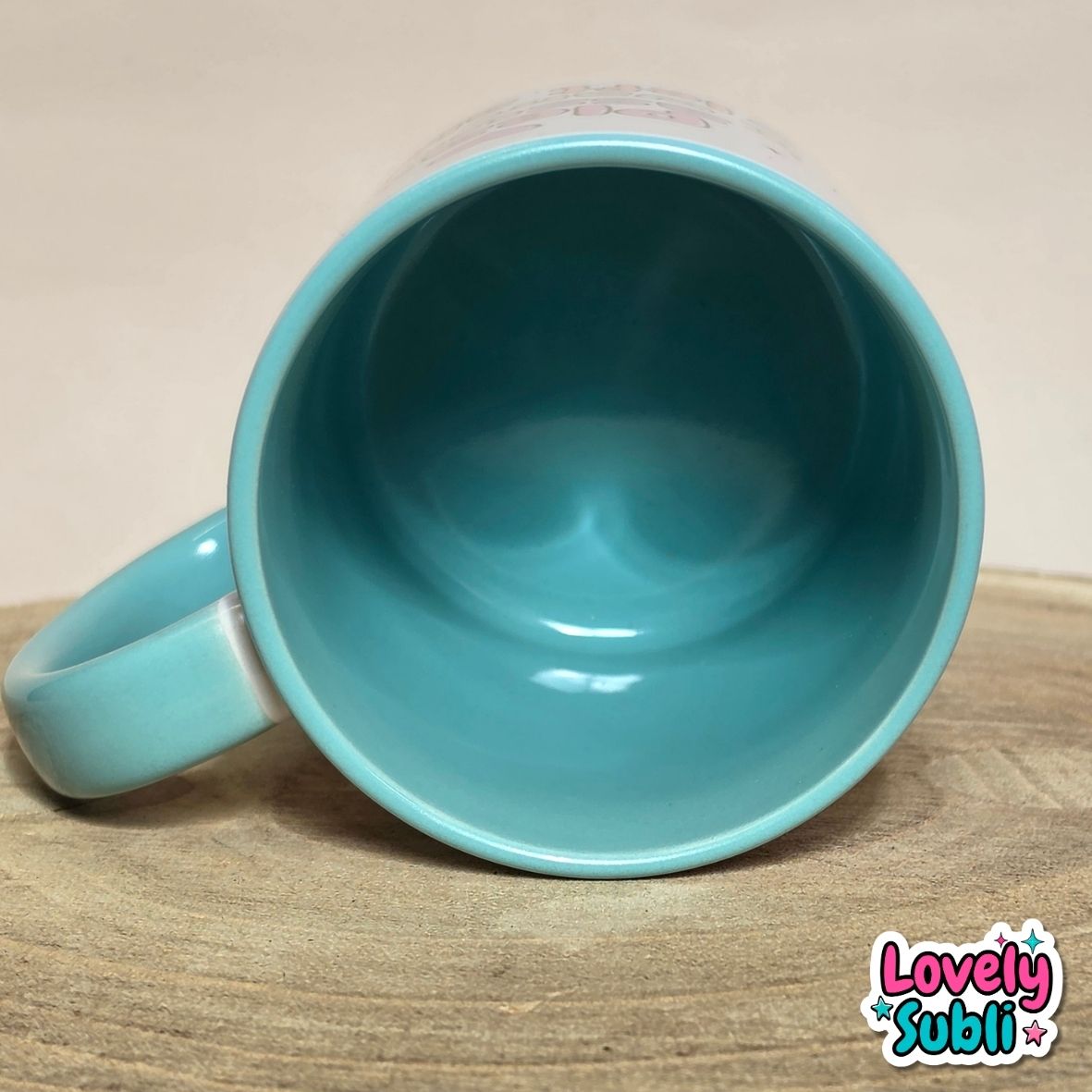 A turquoise ceramic mug shown from a top-down angle, resting on a wooden surface against a neutral beige background. A 'Lovely Subli' brand logo is visible in the bottom right corner.