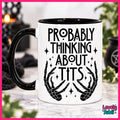 A white ceramic mug with a black handle and black interior, printed with the phrase 'PROBABLY THINKING ABOUT TITS' in black lettering above skeletal hands and sparkles. The mug is photographed on a marble-like surface with a blurred Halloween-themed background.