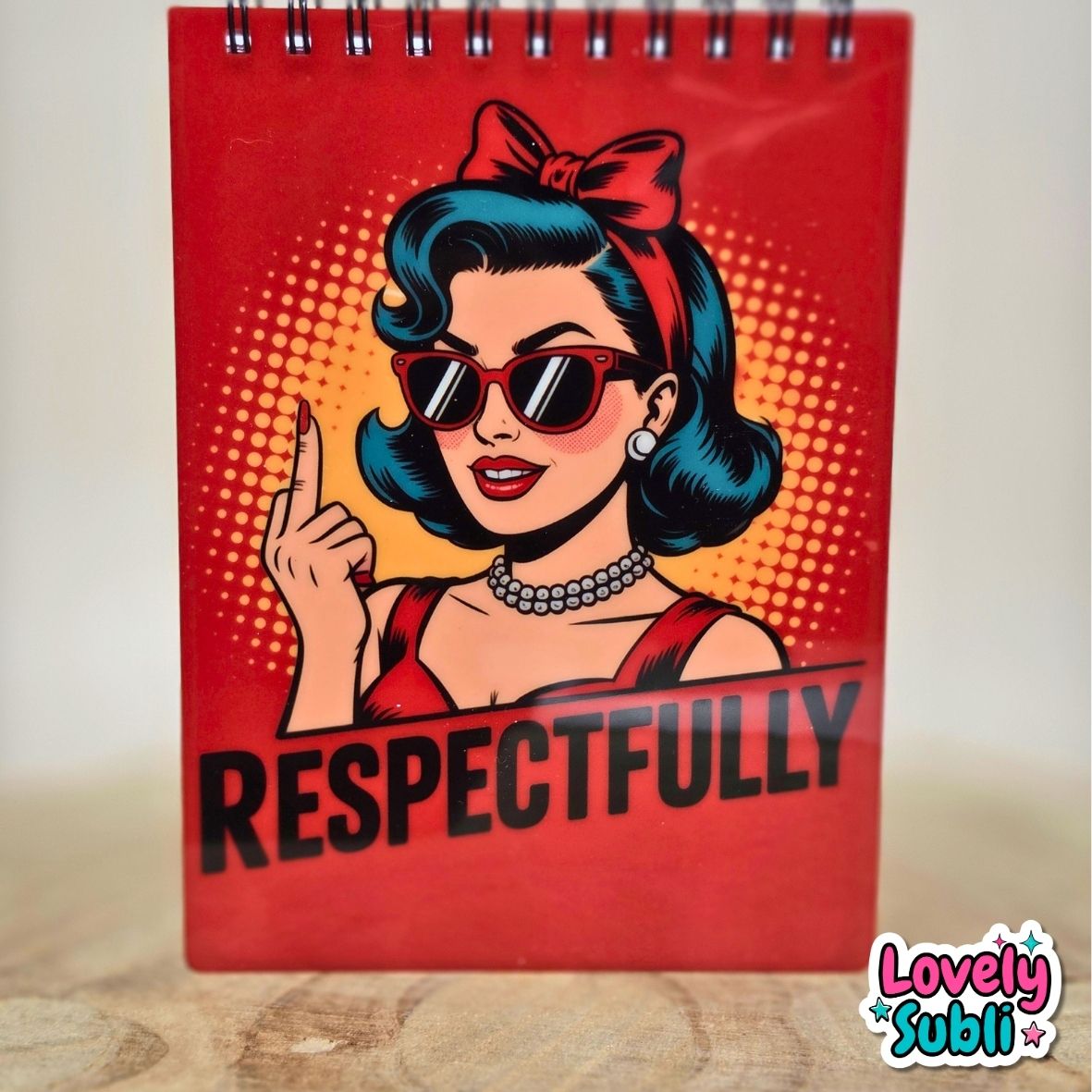 A spiral-bound notebook with a red cover featuring a retro pop-art illustration of a woman with blue hair, sunglasses, and a red bow, with the word 'RESPECTFULLY' printed in bold black letters. The notebook is standing on a light wooden surface against a neutral background.
