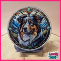 A circular decorative plaque or coaster featuring a stained-glass-style illustration of an Australian Shepherd dog, displayed on a small stand on a wooden surface. The design includes blue, green, brown, black, and white tones.