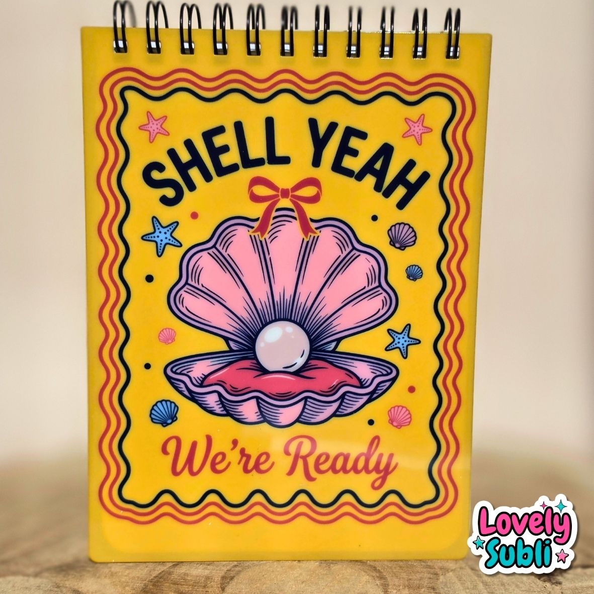 A spiral-bound notebook with a bright yellow cover featuring a pink clamshell illustration and the text 'SHELL YEAH We're Ready'. The notebook is standing upright on a wooden surface against a neutral beige background.