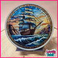 A circular stained glass art piece depicting a sailing ship on blue waves with mountains and a sunset sky, displayed on a small stand. The piece has a white rim and is photographed against a light background with a pink border and a 'Lovely Subli' logo in the bottom right corner.