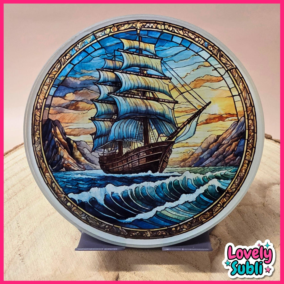 A circular stained glass art piece depicting a sailing ship on blue waves with mountains and a sunset sky, displayed on a small stand. The piece has a white rim and is photographed against a light background with a pink border and a 'Lovely Subli' logo in the bottom right corner.