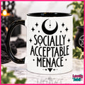 A white ceramic mug with a black handle and black interior, featuring the text 'SOCIALLY ACCEPTABLE MENACE' in black lettering with a crescent moon and stars above. The mug is set on a marble surface with a blurred background containing candles and greenery.