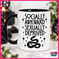 A white ceramic mug with a black handle and black interior, printed with the phrase 'SOCIALLY AWKWARD SEXUALLY DEPRIVED' in black lettering above a black snake illustration with stars. The mug is photographed on a marble-like surface with a softly blurred indoor background and a pink border around the image.
