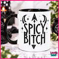 A white ceramic mug with a black handle and black interior, featuring the text 'SPICY BITCH' in a gothic font with crescent moons, stars, and an arrowhead graphic. The mug is displayed on a marble surface with a blurred background containing candles and greenery.
