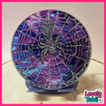 A round decorative plaque or clock face featuring a spiderweb design over a purple and blue iridescent shell-like background, with a metallic spider in the center, displayed on a stand.