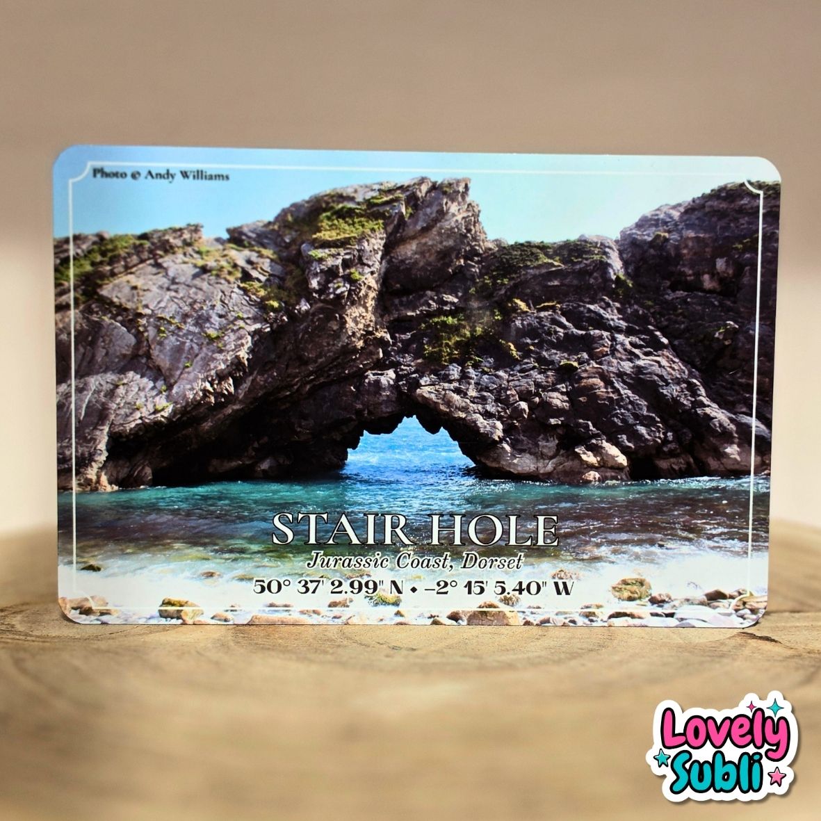 A rectangular printed A6 metal plaque featuring a coastal rock arch scene labeled 'STAIR HOLE Jurassic Coast North,' photographed standing on a tan surface against a beige background. A 'Lovely Subli' logo appears in the bottom-right corner.