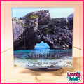 A square coaster featuring a scenic photograph of Stair Hole in Lulworth Cove, Dorset, displayed on a small black stand. The coaster is set against a light wood background with a pink border, and a 'Lovely Subli' logo is visible in the bottom right corner.