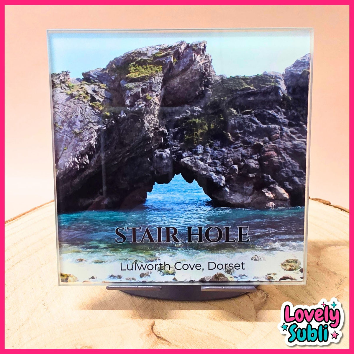 A square coaster featuring a scenic photograph of Stair Hole in Lulworth Cove, Dorset, displayed on a small black stand. The coaster is set against a light wood background with a pink border, and a 'Lovely Subli' logo is visible in the bottom right corner.
