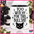 A white ceramic mug with a black handle and black interior, printed with the text 'TOO WITCHY FOR THIS BULLSHIT' in black lettering, surrounded by black stars and a black cat face with a crescent moon. The mug is shown on a marble-like surface with a blurred Halloween-themed background.
