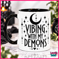A white ceramic mug with a black handle and black interior, printed with the phrase 'VIBING WITH MY DEMONS' in black lettering alongside a crescent moon and star motifs. The mug is shown on a marble-like surface with a blurred gothic-themed background.