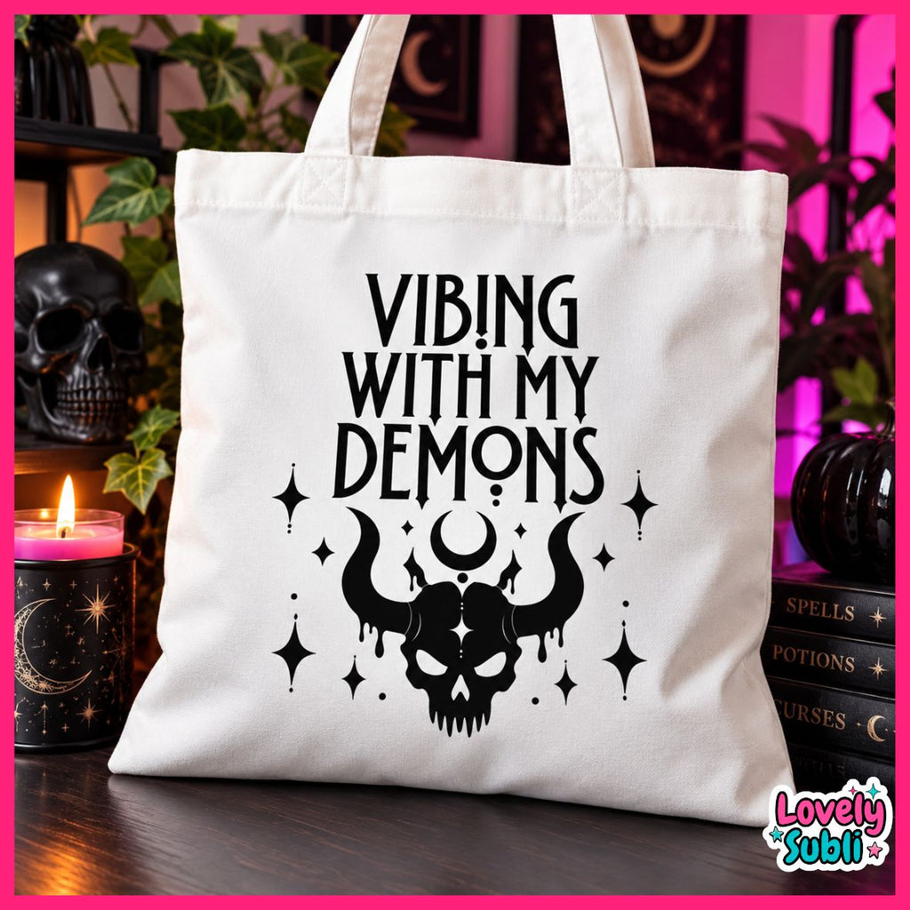 A white canvas tote bag with black printed text reading 'VIBING WITH MY DEMONS' and a horned skull graphic, displayed on a dark tabletop with candles, books, and plants in a styled indoor setting.