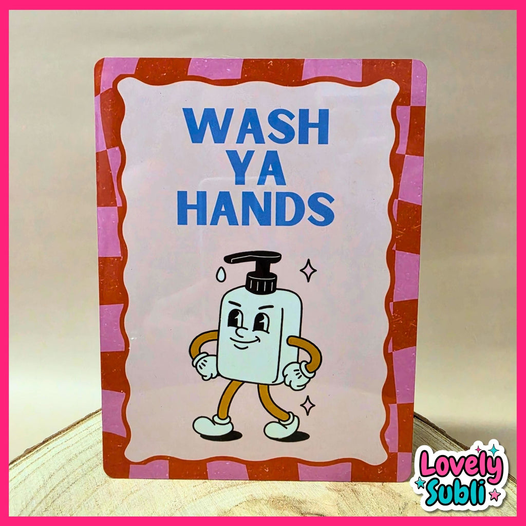 A rectangular handwashing sign with the words 'WASH YA HANDS' in blue text and a cartoon soap bottle character. The sign has a pink and orange checkered border and stands on a light wooden surface against a beige background.