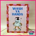 A rectangular handwashing sign with the words 'WASH YA HANDS' in blue text and a cartoon soap bottle character. The sign has a pink and orange checkered border and stands on a light wooden surface against a beige background.