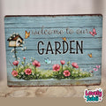 A rectangular rustic wooden sign with a distressed light blue wood-plank background, featuring the words 'welcome to our GARDEN' in black script and block lettering. The sign is decorated with pink flowers, green leaves, a small birdhouse, butterflies, and white blossoms.