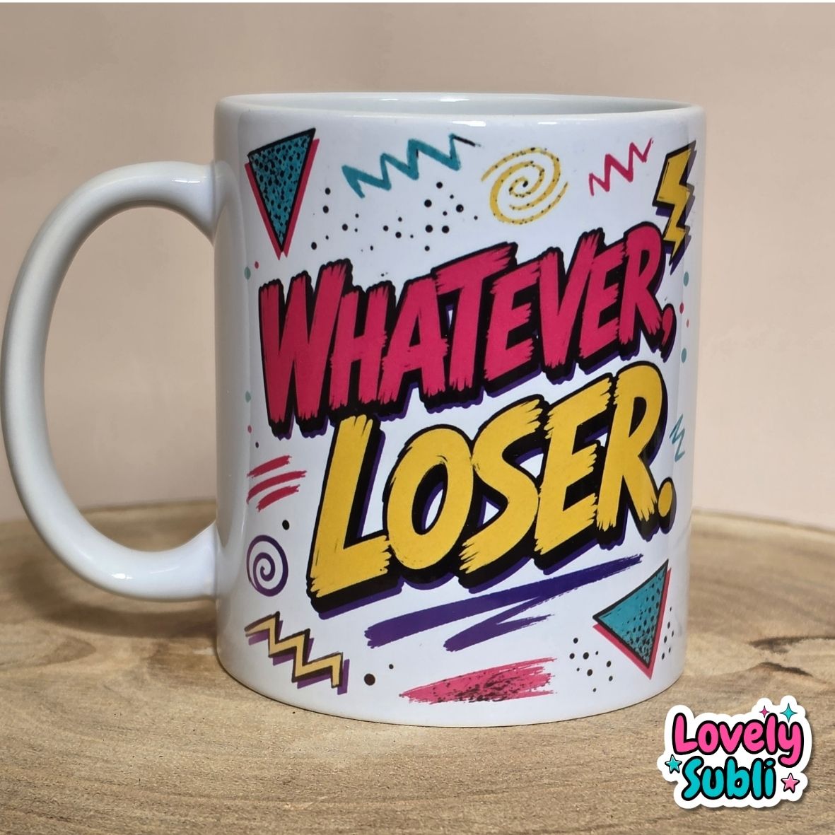 A white ceramic mug with a colorful retro graphic that reads 'WHATEVER, LOSER.' in pink and yellow bubble letters, surrounded by doodle-style shapes, shown on a wooden surface against a beige background.