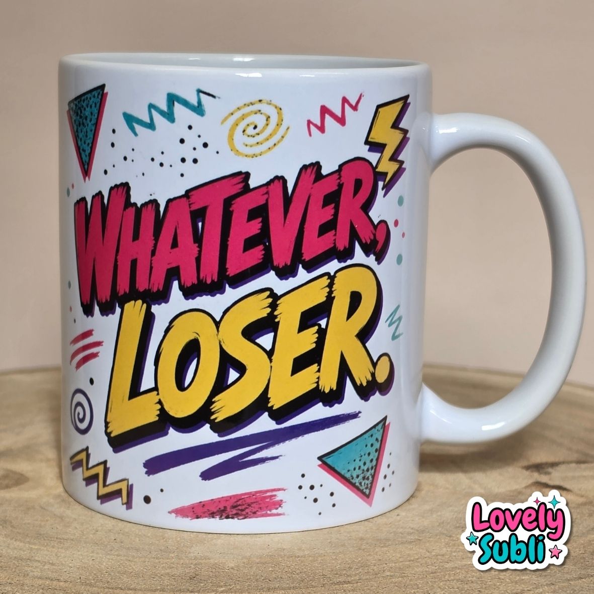 A white ceramic mug featuring the phrase 'WHATEVER, LOSER.' in bold pink and yellow retro comic-style lettering, surrounded by colorful geometric doodles, displayed on a wooden surface against a beige background.