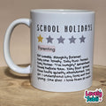 A white ceramic mug printed with the text 'SCHOOL HOLIDAYS' and a humorous parenting definition, sitting on a wooden surface against a beige background.