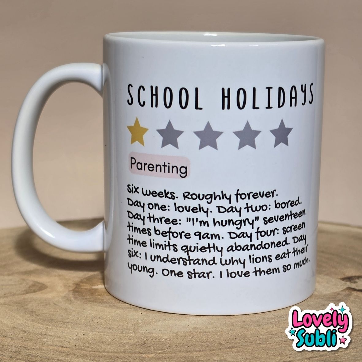 A white ceramic mug printed with the text 'SCHOOL HOLIDAYS' and a humorous parenting definition, sitting on a wooden surface against a beige background.