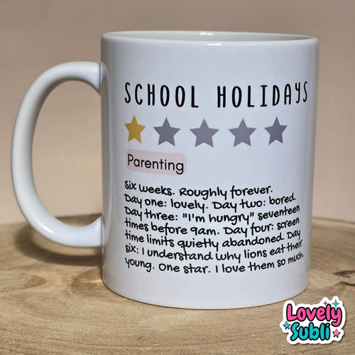 A white ceramic mug printed with the text 'SCHOOL HOLIDAYS' and a humorous parenting definition, sitting on a wooden surface against a beige background.