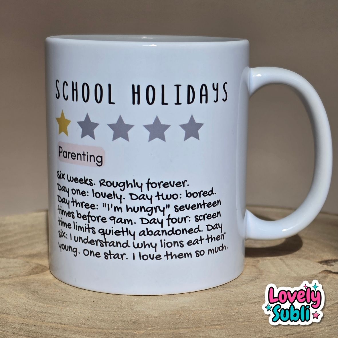 A white ceramic mug printed with the text 'SCHOOL HOLIDAYS' and a humorous parenting definition with a one-star rating, shown on a wooden surface with a neutral background.
