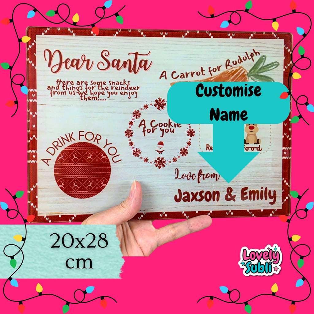 A hand holds a personalized Christmas letter board or plaque against a bright pink background with decorative string lights. The board has a rustic white wood texture with red borders, holiday-themed text including 'Dear Santa', 'A Carrot for Rudolph', 'A Cookie for you', and 'Love from Jaxson & Emily', plus a turquoise arrow labeled 'Customise Name'.