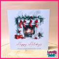 A square Christmas greeting card featuring a watercolor illustration of a fireplace with red stockings, green garland, gifts, and the text 'Happy Holidays' in red script. The card is photographed standing on a light wood surface against a soft beige background, with a pink border and a 'Lovely Subli' logo in the lower right corner.