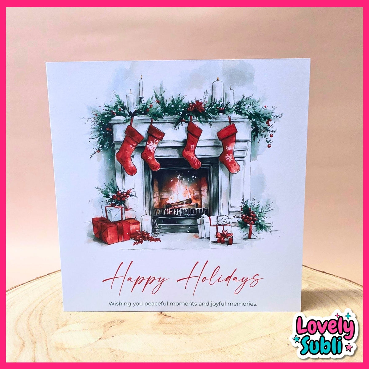 A square Christmas greeting card featuring a watercolor illustration of a fireplace with red stockings, green garland, gifts, and the text 'Happy Holidays' in red script. The card is photographed standing on a light wood surface against a soft beige background, with a pink border and a 'Lovely Subli' logo in the lower right corner.